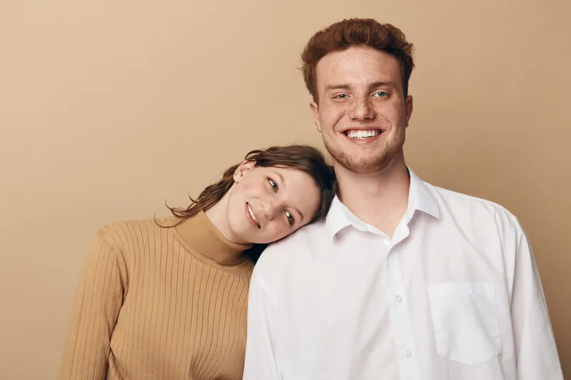 Smiling young man in white shirt and woman in brown sweater lean on his shoulder, showing affection and happiness. Neutral background creates warm and cozy atmosphere of friendship and love.