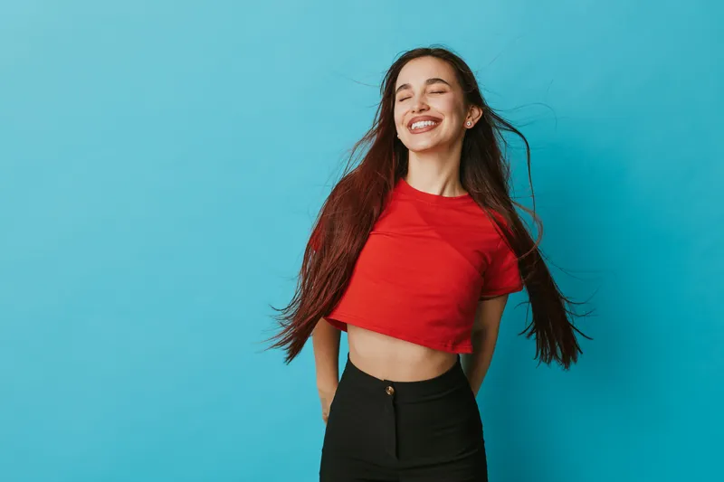 Young woman with long hair wearing red crop top and black pants smiling with closed eyes against blue background. Female model posing with carefree expression and natural beauty.