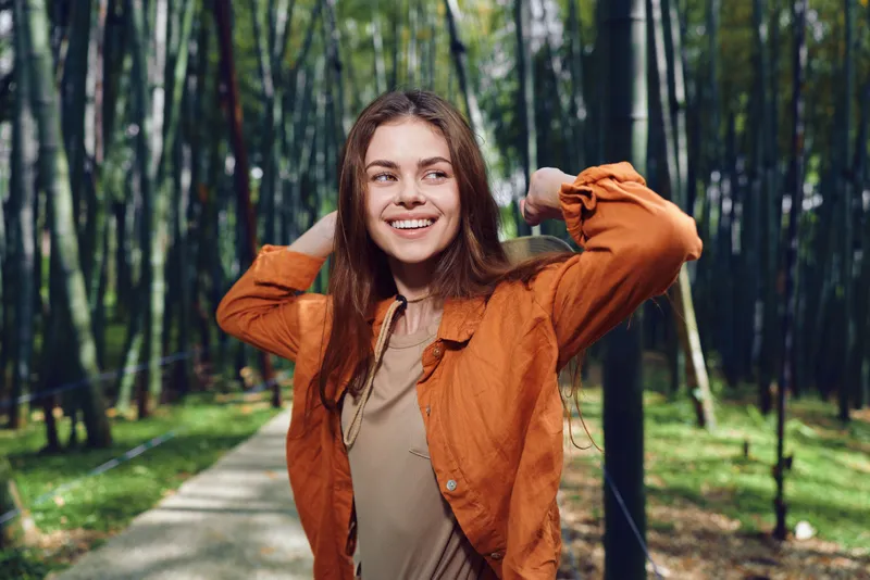 woman smile portrait in park with bamboo path, young female in orange jacket posing with hands behind head, natural sunlight, casual happy expression and relaxed outdoor mood
