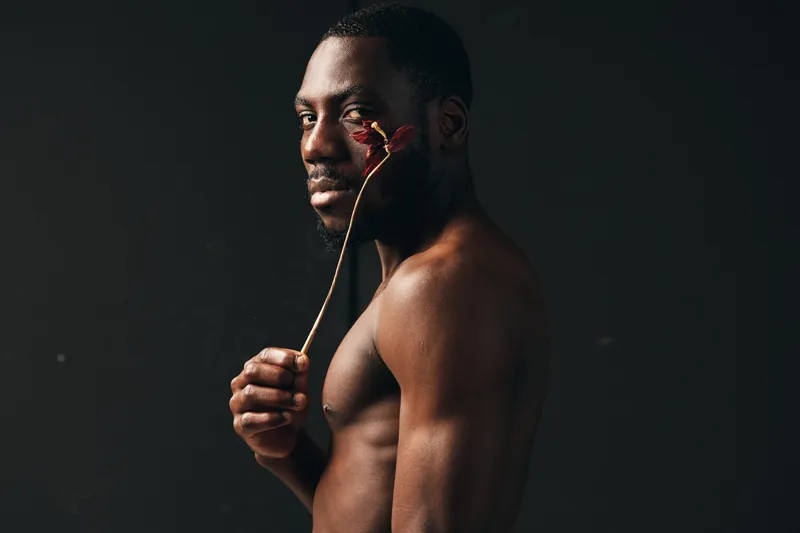 Creative conceptual portrait of African American man with bare torso holding dried flower near face against dark background. Artistic visual style with focus on expression and texture.