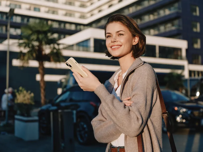A cheerful professional woman stands outside a modern office building, texting on a smartphone. She smiles warmly, conveying a positive and engaging business atmosphere.