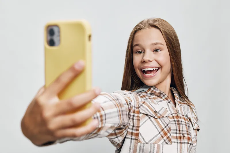 Smiling girl taking selfie with yellow phone in casual plaid shirt, happy teenager capturing photo on smartphone against plain light background indoors.