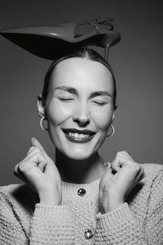 Happy white woman with fair skin smiling with closed eyes holds fists near face, balancing a high heel shoe on her head. Concept for joy, fashion, and fun in black and white.