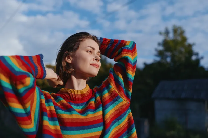 Young woman in a rainbow striped sweater outdoors in a rural setting, smiling with arms raised, short brown hair and casual outfit, warm sunlight creating a peaceful lifestyle portrait.