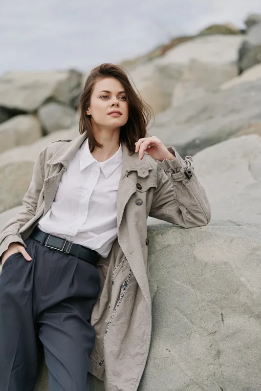 Young woman in stylish coat and white shirt leans on large rocks outdoors. Casual fashion portrait with natural makeup and relaxed confident pose in overcast weather.