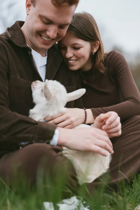 Cheerful couple outdoors, tenderly cradling a kid goat, warm smiles, sunlit grass, soft breeze, natural colors, intimate moment, relaxed pose, rustic vibe, lifestyle scene with animals