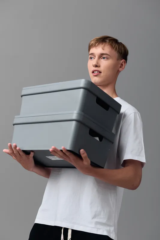 Young man in a white shirt holding a stack of plastic storage boxes, illustrating organization, storage, and home shelving in a clean studio setting today