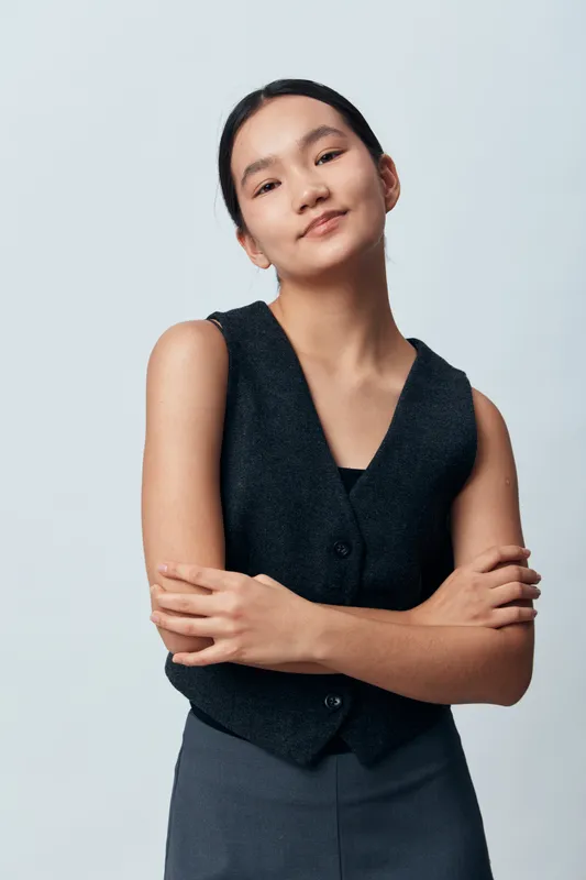 Young confident woman with crossed arms standing against plain background wearing dark vest and pants, looking at camera with slight smile and relaxed posture.