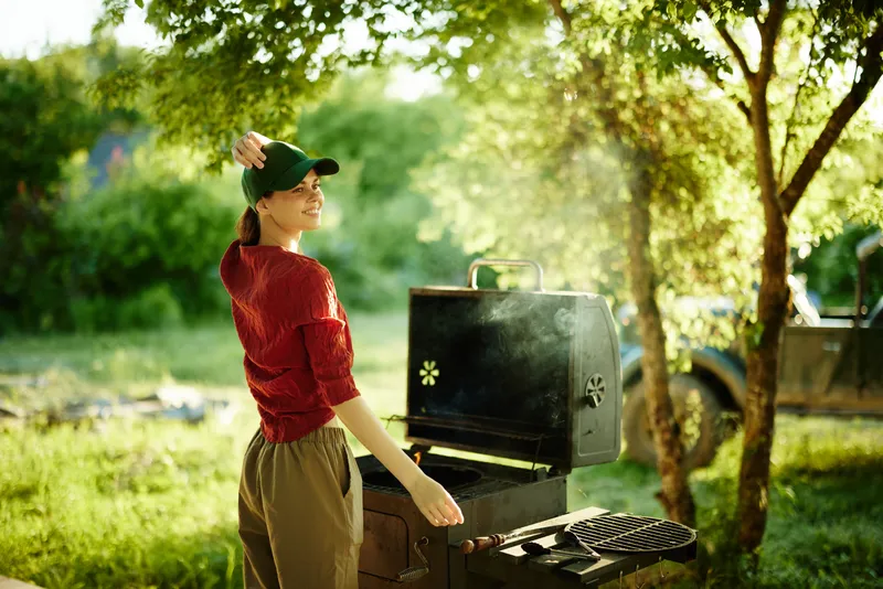 Young woman grilling outdoors, showcasing joy and relaxation in a lush garden setting with green foliage and smoke from a barbecue grill