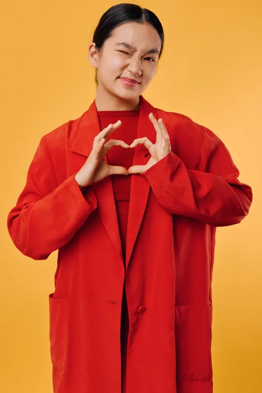 Young woman in red coat on yellow background making heart shape with hands, smiling with eyes closed for love, affection and positive emotion in studio portrait.