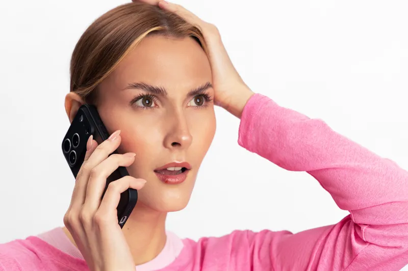 Concerned young woman in pink sweater talking on smartphone with worried expression holding head. Isolated on white background. Concept for stress, communication, and emotional reaction.