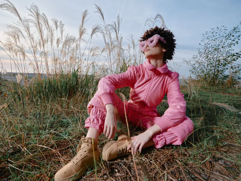 Outdoor fashion scene with a model in a pink jumpsuit, boots and a stylish mask, posing in tall grass under soft sunlight for a relaxed lifestyle mood.