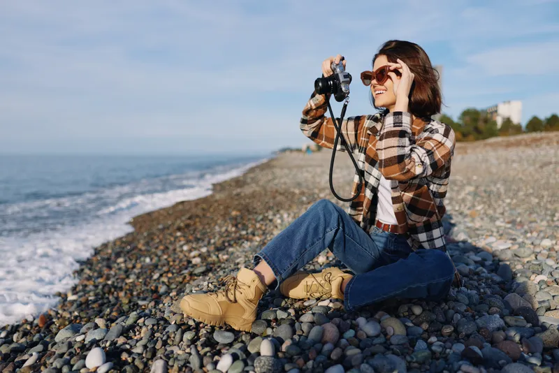 Woman photographer on a pebble beach, aiming a camera toward the sea, wearing plaid shirt, jeans and boots, enjoying outdoor photography by the coast in daylight.
