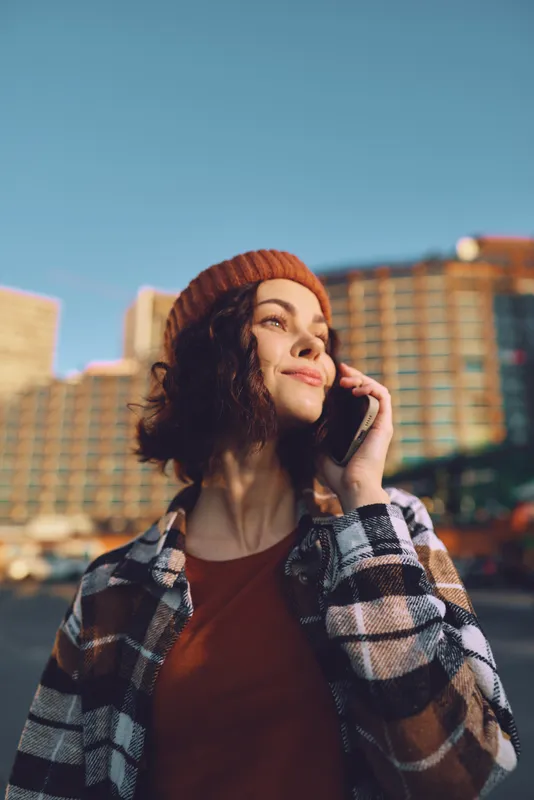Woman with phone and smile in urban street at golden hour, candid authenticity and mindful living mood. Beanie and sunlight, emotional storytelling portrait with modern city aesthetic.