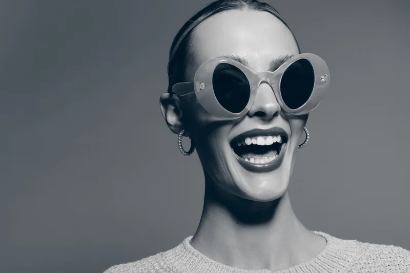 Happy white woman with fair skin wearing round sunglasses smiles with joy on neutral grey background. Concept of fashion, style, and positive emotions.