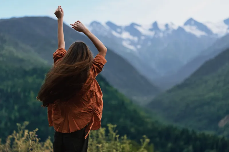 Woman with arms raised stands in a vast mountains landscape, celebrating freedom and a deep connection with nature in an expansive outdoor setting beneath clear skies and rugged peaks.