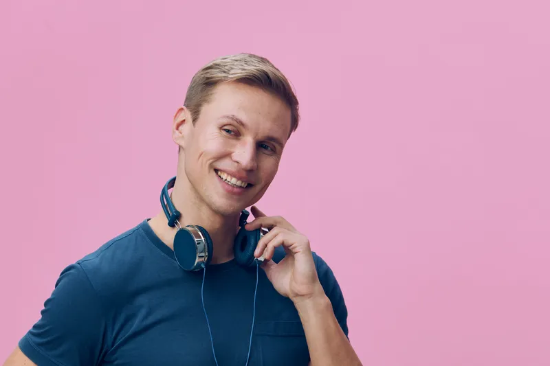 Smiling young caucasian man wearing casual blue t-shirt with headphones around neck posing on pink background studio portrait. Lifestyle, cheerful, happy, youth, fashion, music concept