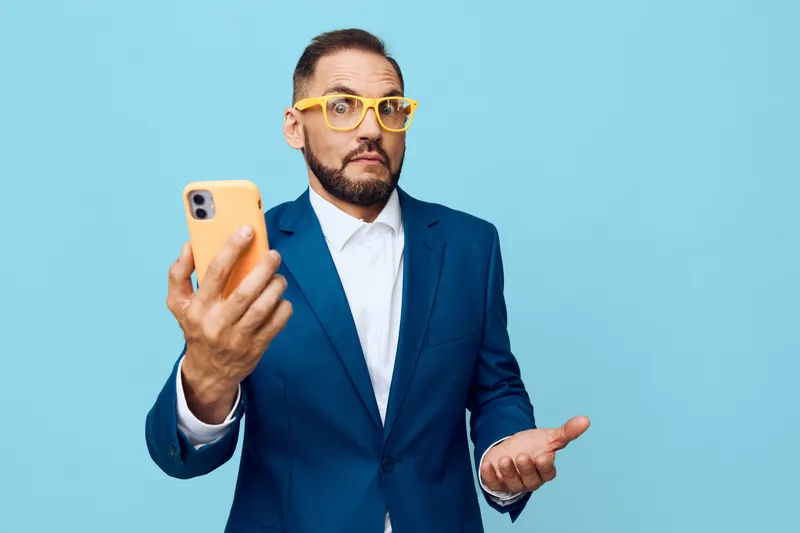 A confident male business professional stands against a bright blue background, holding a smartphone. His posture and gaze convey curiosity, focus, and readiness for action.