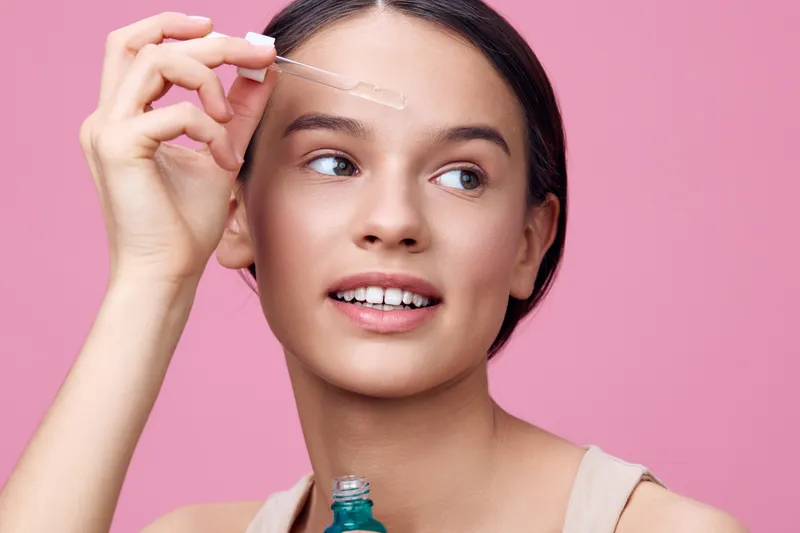 Young woman applying facial serum with a dropper, showcasing healthy skin and beauty care against a pink background Keywords skincare, beauty, serum, healthy skin, feminine