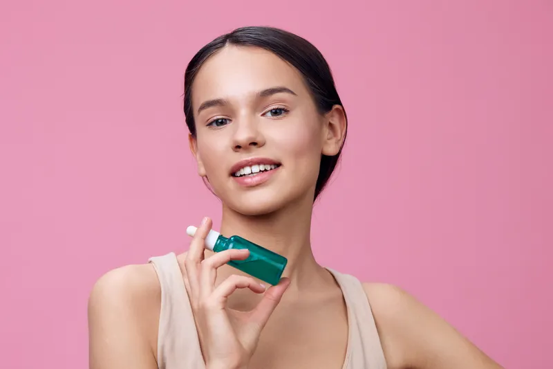 Young woman holding a green skincare bottle, smiling confidently against a pink background, promoting healthy skincare and beauty concepts