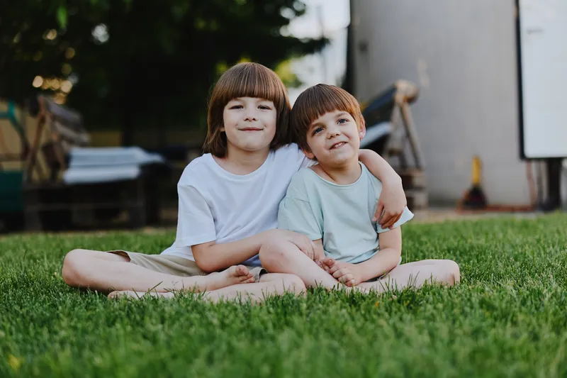 Two smiling boys sitting together on the grass, enjoying their time outdoors with a warm, sunny atmosphere and playful expressions.