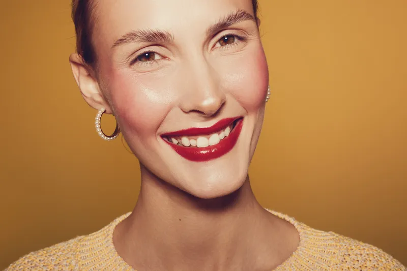 Close-up portrait of a smiling woman with red lipstick and hoop earrings on warm brown background. Concept of beauty, happiness, makeup, and natural skin glow in studio lighting.