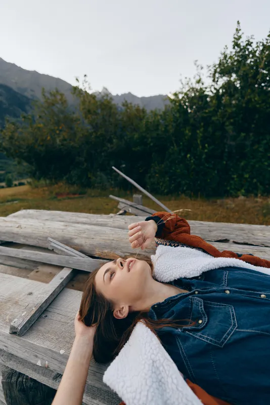 Woman portrait relaxed lying on wooden platform in nature near mountains and meadow, wearing jacket and denim shirt, outdoors leisure with peaceful expression and scenic countryside view.