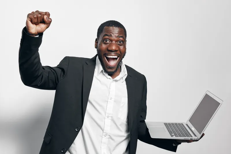 African man with dark skin in black suit holding laptop and celebrating success with raised fist and wide smile on white background. Excited businessman in formal wear.