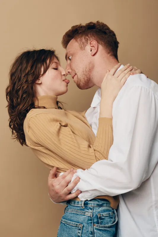 Young couple in casual clothes embraces closely, about to kiss. Intimate moment captured in warm studio lighting on beige background. Concept of love, romance, and connection.