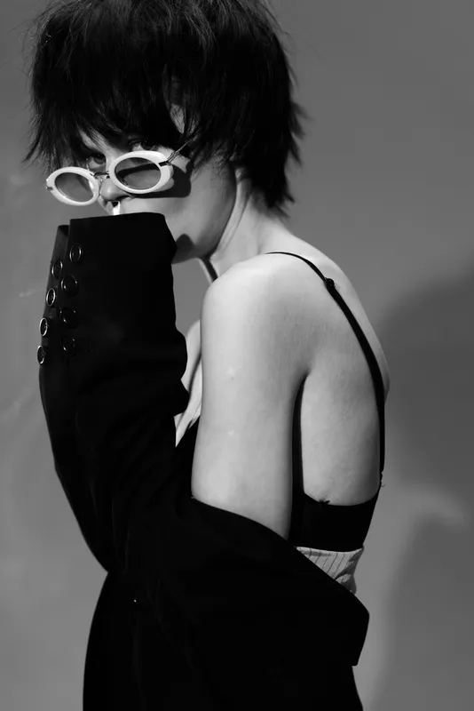 Black and white portrait of a stylish woman wearing white sunglasses and black clothing with moody expression and edgy hairstyle.