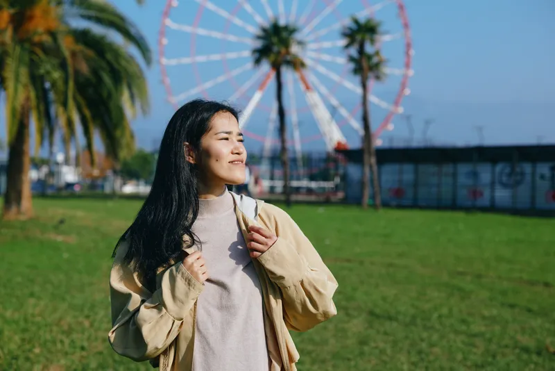 Woman smiling in a park on a sunny day, ferris wheel in the background, casual jacket and sweater, outdoor leisure moment with greenery and daylight, calm atmosphere and breeze.