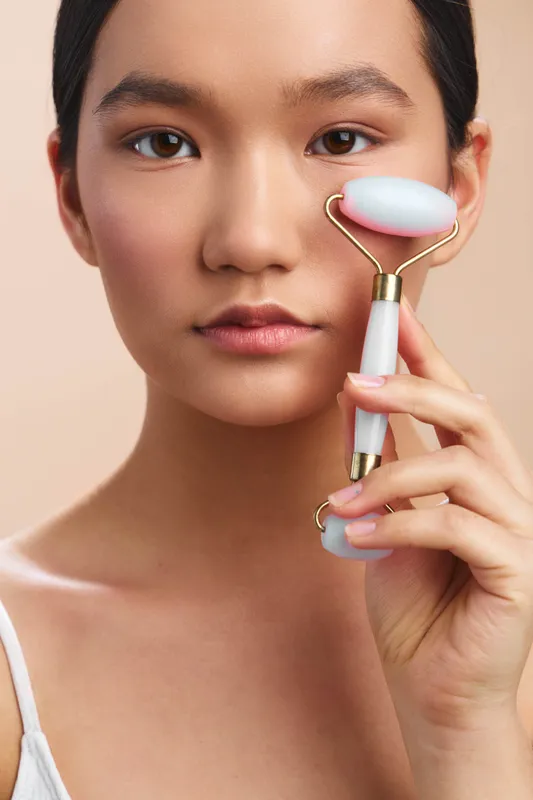 asian woman uses a facial roller in a calm personal care setting, focusing on skincare routine and relaxation. she smiles softly, creating a peaceful and fresh atmosphere with clean light.