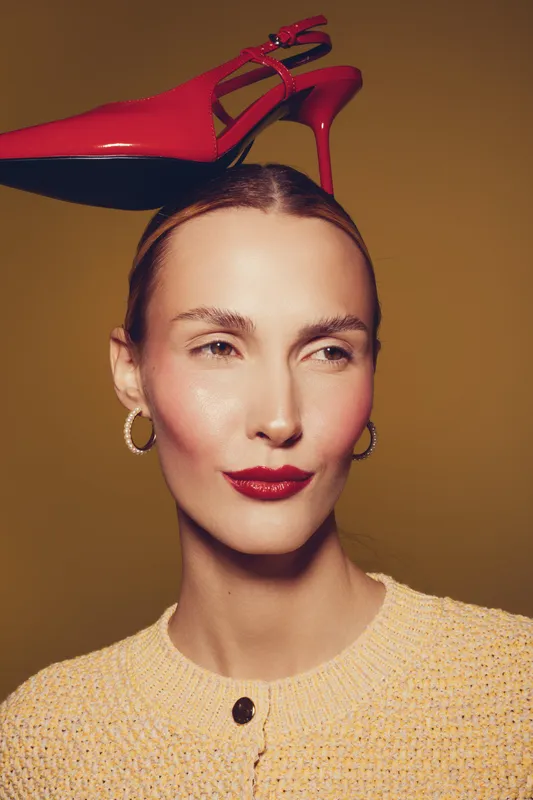 Woman with red high heel shoe balanced on head wears yellow sweater and hoop earrings. Studio portrait with warm lighting and brown background. Concept of fashion and creativity.