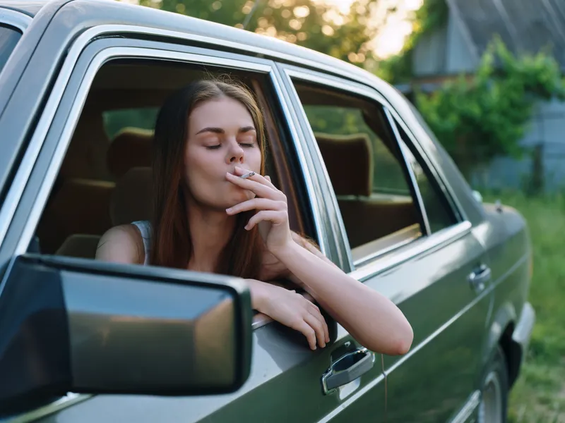 Young woman enjoying a cigarette while sitting in a car, capturing a moment of relaxation and contemplation in a natural outdoor setting.