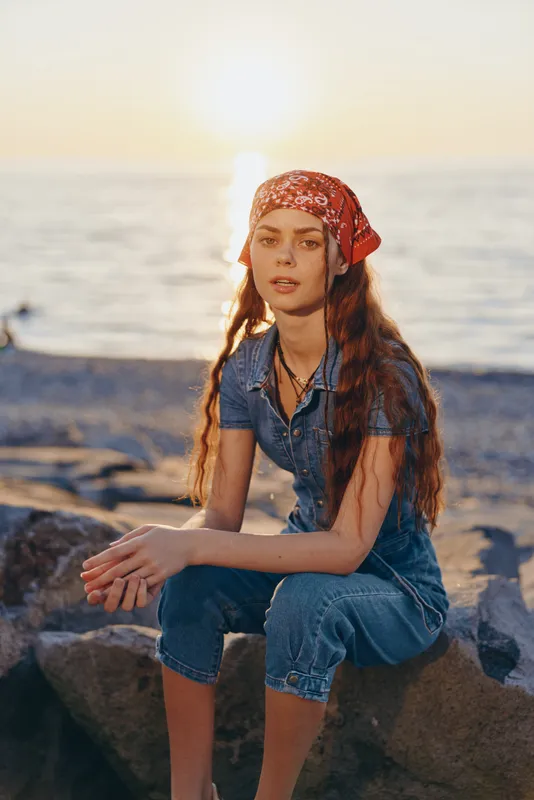 lifestyle woman in modern boho-western denim street style with red bandana sitting by the beach during golden hour, film color vintage warm sunlit mood