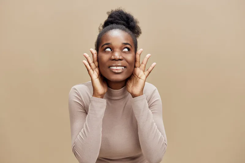 Happy young African woman smiling and posing against a neutral background, hands on ears illustrating a joyful expression, perfect for wellness and positive emotions
