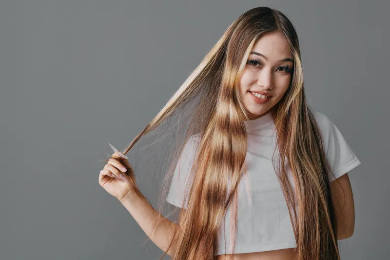 Young woman with long straight hair smiling while holding strands in hand wearing a white t shirt against plain background for beauty and fashion concept.