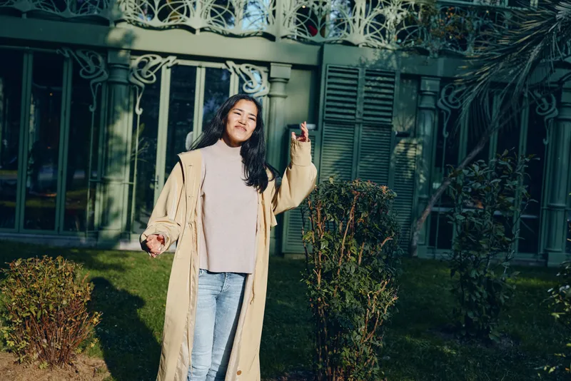 Smiling woman posing outdoors in a long beige coat over a knit sweater and jeans, sunny garden setting with hedges and gate, casual fashion lifestyle shot emphasizing style, warmth and confidence