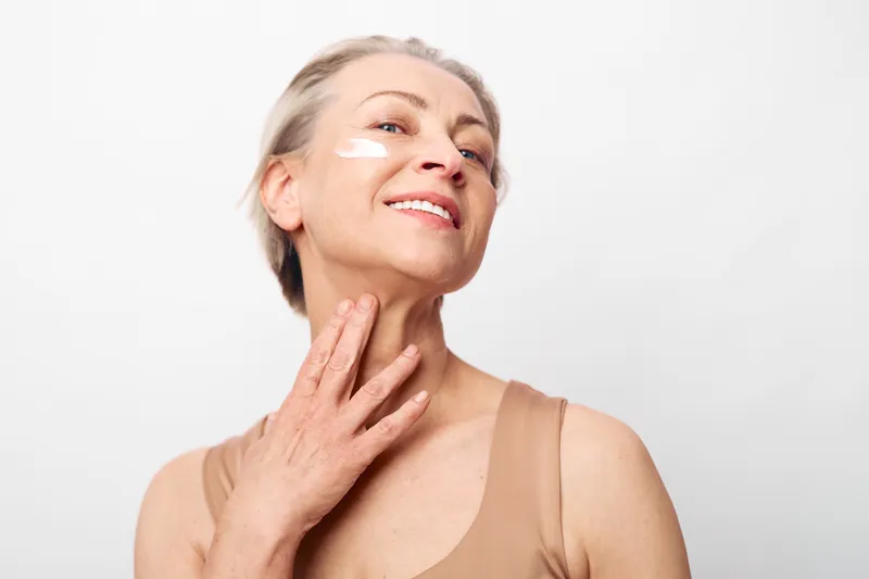 Mature woman with light skin applies skincare cream on her face and neck. She smiles gently in a bright studio with soft lighting. Concept of anti aging, beauty, and self care.