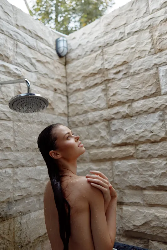 woman showering outdoors with long wet hair, stone wall background, natural light, peaceful mood, spa wellness concept in warm tones