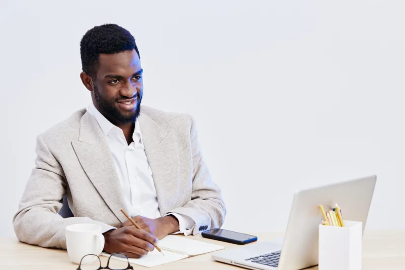 Professional young African American man in formal blazer and white shirt working at desk with laptop, notebook, coffee mug, and pen in modern office environment. Confident business professional, works
