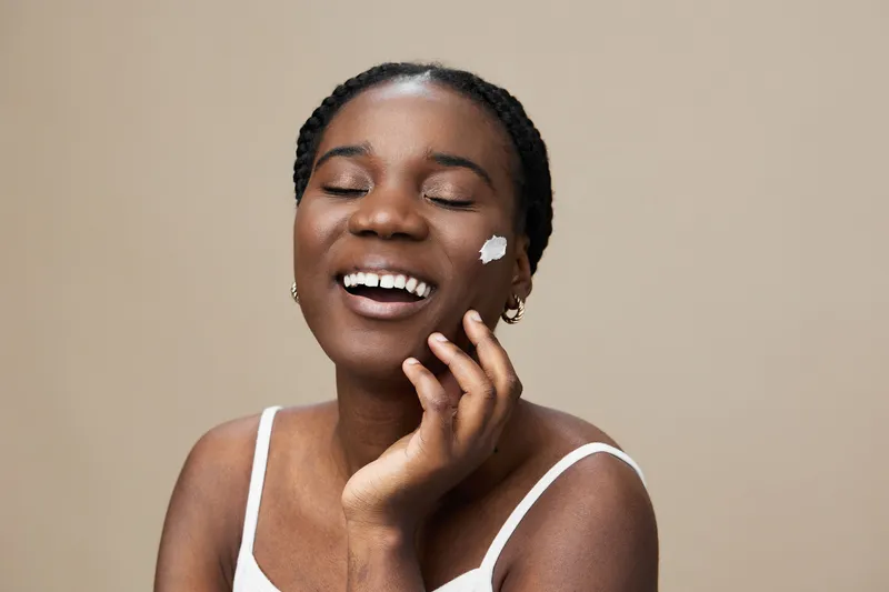 Joyful Black woman applying skincare on her face, smiling brightly against a neutral background, showcasing natural beauty and self care