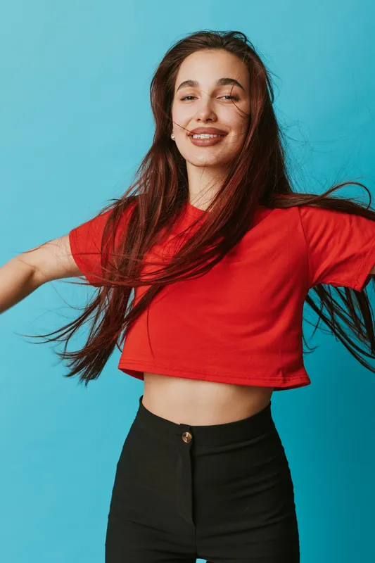 Young woman smiling and dancing with long hair wearing red crop top and black pants against blue background. Casual style, happiness and movement for lifestyle and youth.