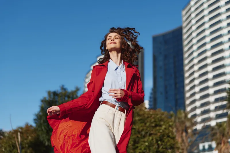 A young woman in a red coat walks through a sunny urban plaza, hair and coat fluttering in the breeze, capturing confident street style.