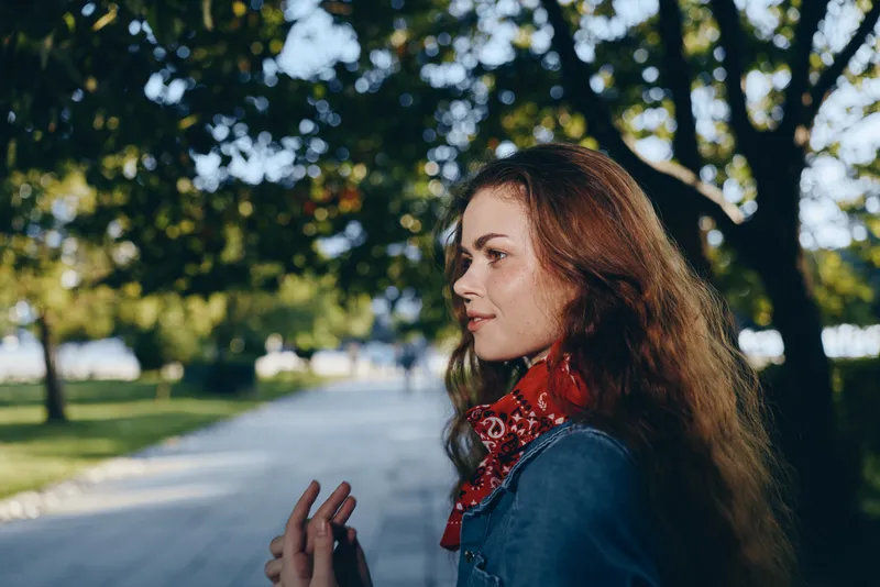 Profile of a red-haired woman wearing a denim jacket and red scarf outdoors with green leafy background under natural sunlight during daytime.