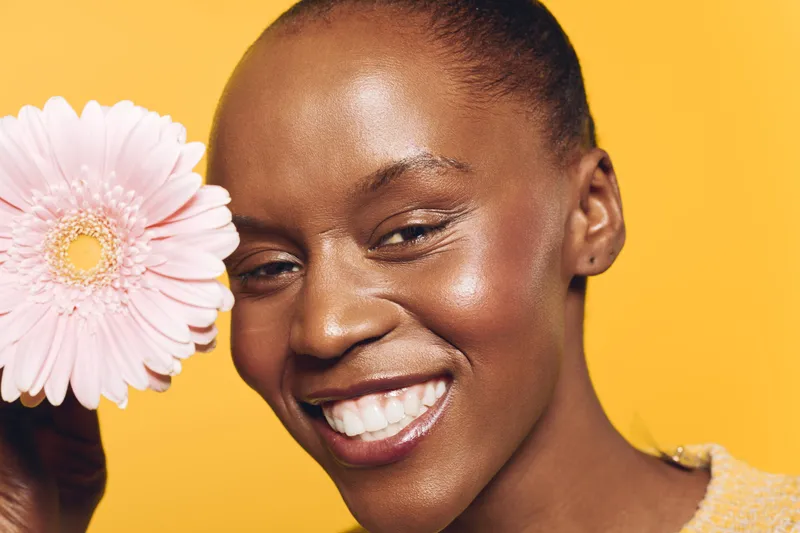 Close-up of happy African woman with dark skin smiling brightly while holding pink flower. Yellow background highlights natural beauty, skincare, and positive mood in studio lighting.