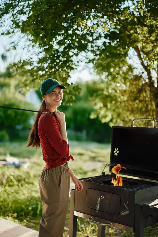 Young woman grilling outdoors with a joyful expression, wearing casual clothing and a cap by a green nature background Perfect for seasonal cooking concepts and outdoor activities