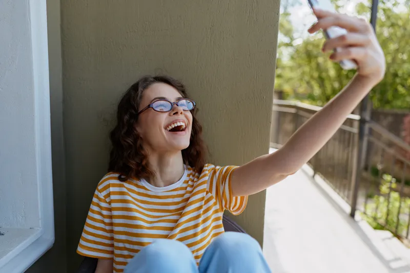 Smiling woman wearing a striped shirt takes a selfie on a balcony, natural daylight on a sunny day, casual home lifestyle, relaxed mood, joyful moment captured outdoors near a railing.