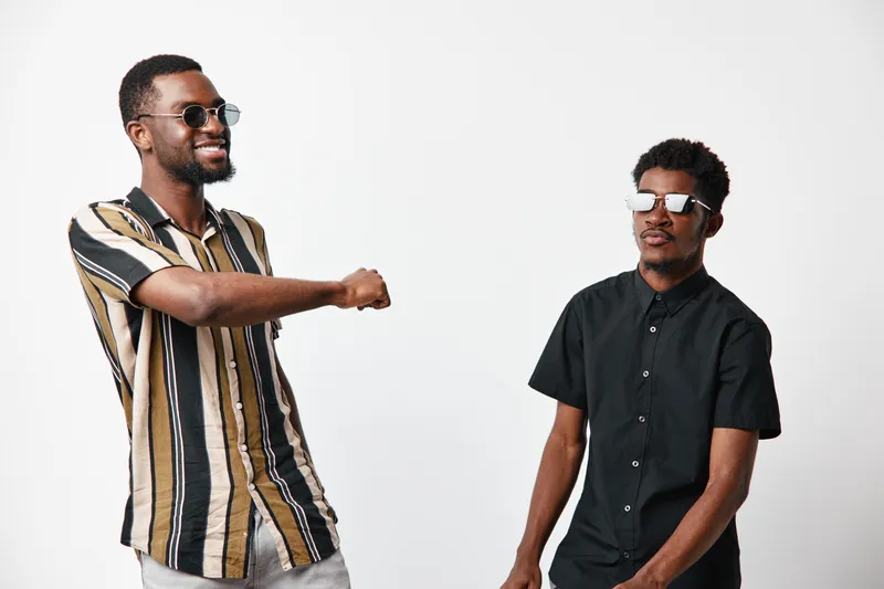 Two African American men with dark skin wearing sunglasses greeting each other with elbow bump on white background. Casual style, friendship and social interaction concept.