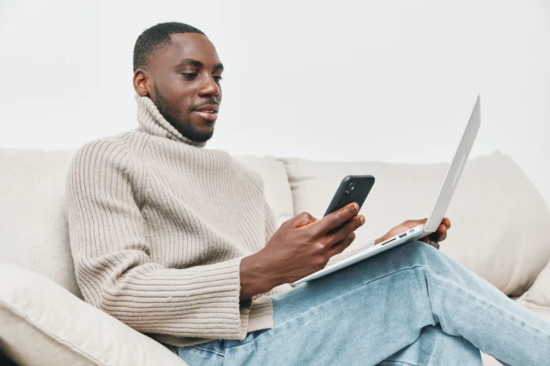 A young Black man in a cozy cream turtleneck sweater enjoys using his smartphone while sitting on a sofa with a laptop, showcasing modern lifestyle and technology